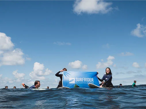 alt="Group of ocean activists floating in the sea holding a Surfrider Foundation Europe banner under a blue sky."