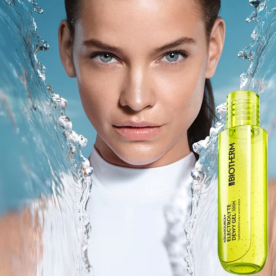 alt="Woman with glowing, hydrated skin surrounded by water splashes, alongside a neon yellow-green transparent gel bottle against a blue background."