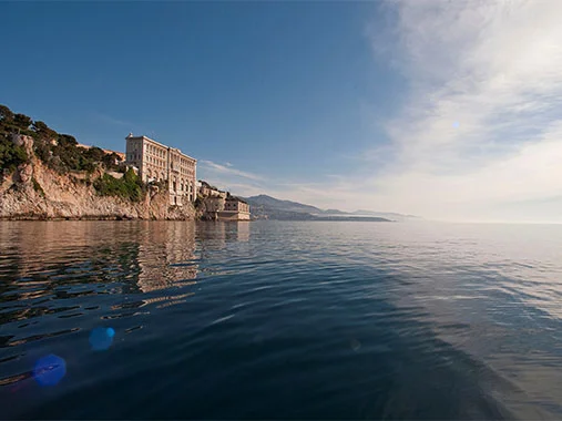 alt="Panoramic sea-level view of the grand stone Oceanographic Museum of Monaco built into a rocky clifftop, its façade reflected in the still blue Mediterranean waters on a clear sunny day."